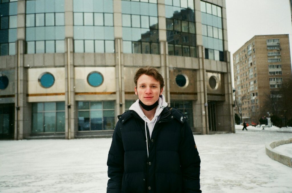 Smiling young man wearing mask outside a modern building on a snowy winter day.